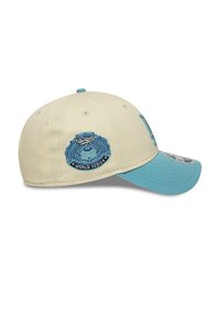 Gorra de béisbol beige con visera azul, que presenta un logotipo bordado de los Dodgers y un parche de la Serie Mundial en el lateral, fabricada con material de algodón.