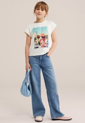 Jong meisje staat met één hand in de zak, draagt een wit T-shirt met flamingo-zonnebril, wijd uitlopende spijkerbroek, roze schoenen en houdt een blauwe handtas vast.