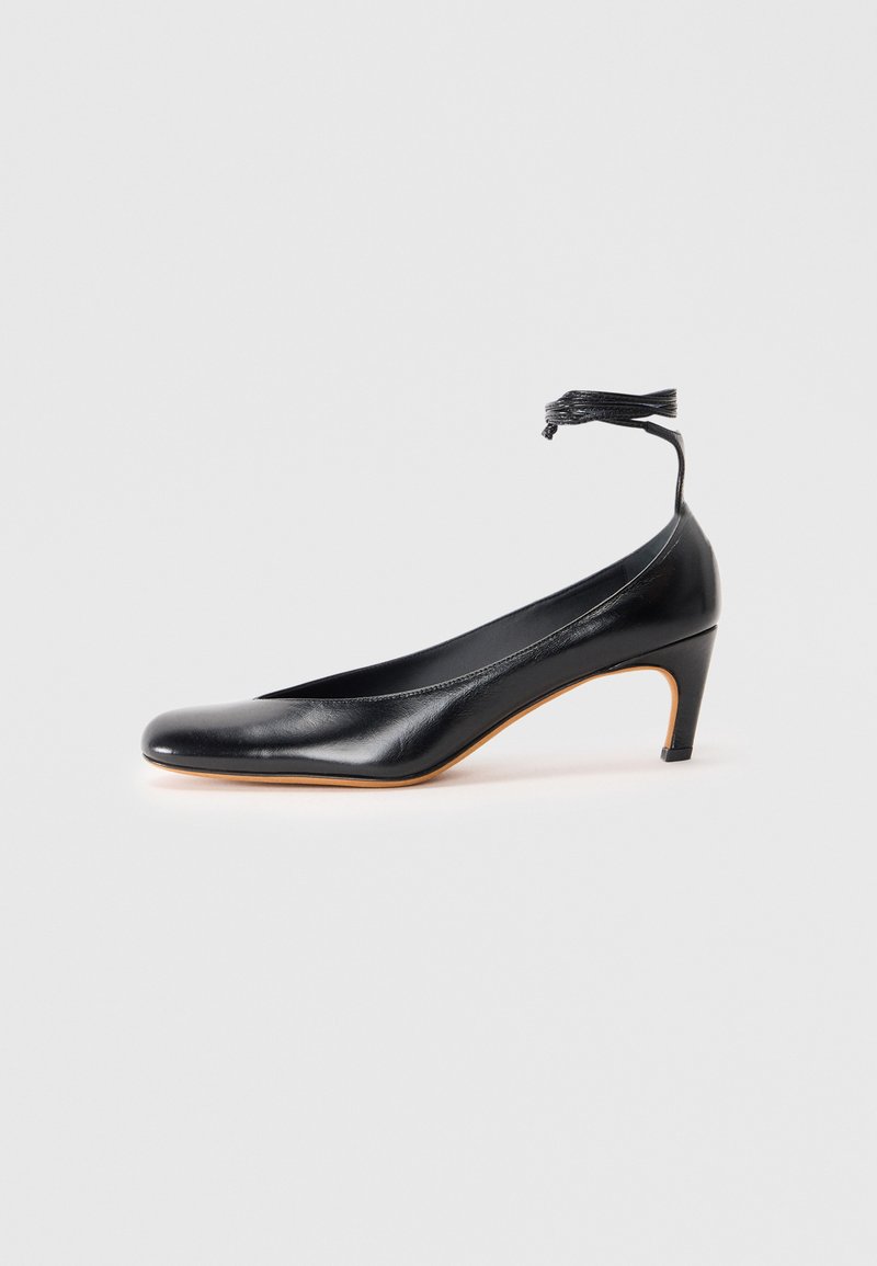 Pantofii negri din piele cu vârful ascuțit, toc subțire și detaliu cu curea la gleznă, având un accent din ciucure. Textură netedă și design elegant.