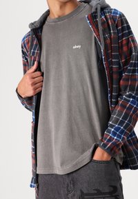 Flanellen jas met rood, blauw en zwart geruit patroon, eronder een grijs katoenen T-shirt, en grijze spijkerbroek met een subtiele print.