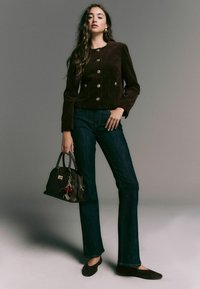 Brown corduroy jacket with gold buttons, flared dark jeans, and a black handbag with red accents. Model stands in a neutral backdrop.