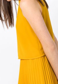 Top amarillo sin mangas con falda plisada, hecha de una tela suave. El diseño presenta un dobladillo en capas y una textura suave para mayor confort.