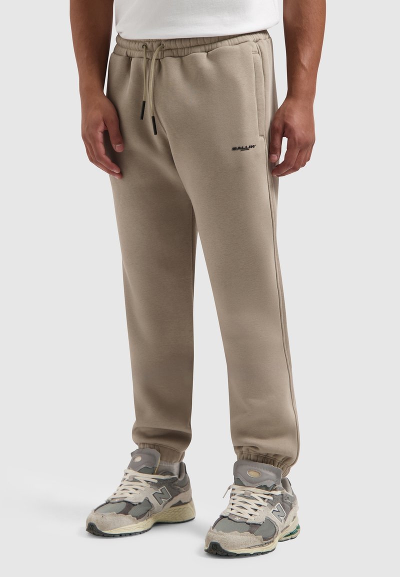 Beige joggingbroek van zacht materiaal, met een elastische tailleband, zijzakken en een klein zwart logo. Gepaard met grijze New Balance sneakers.