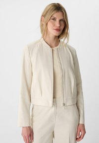 Kremowa kurtka bomberka z okrągłym dekoltem, z zamkiem błyskawicznym z przodu i kieszeniami po bokach. Wykonana z gładkiego, lekkiego materiału.