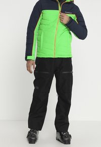 Homme portant une veste verte et marine, un pantalon d'extérieur noir et des chaussures de ski noires, debout devant un fond uni.