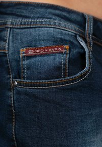 I jeans in denim blu scuro presentano un'etichetta rossa cucita sul taschino posteriore con filo dorato e cuciture arancioni a contrasto lungo le cuciture.
