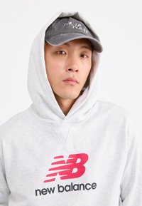 Junger Mann, der einen hellgrauen New Balance-Hoodie mit hochgezogenem Kapuzen und einer dunkelgrauen Baseballmütze trägt, auf der "Sur La Plage" steht, vor einem weißen Hintergrund.