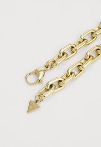 Collana in oro con maglie spesse, chiusura a moschettone e un'etichetta triangolare incisa con "GUESS," che mette in mostra una finitura lucida e brillante.