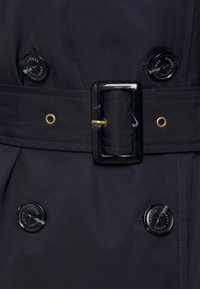 Marineblauwe trenchcoat met een tailleband, voorzien van glanzend zwarte knopen en een rechthoekige gespsluiting. Het materiaal oogt glad met een subtiele textuur.