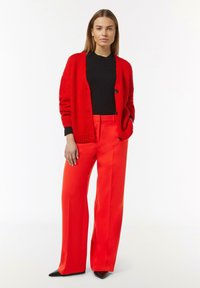 Red textured cardigan over a black top, paired with high-waisted, bright red wide-leg trousers and pointed black heels.