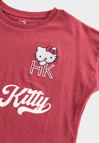 Czerwona bawełniana koszulka z haftowanym wzorem Hello Kitty z kokardką oraz literami "HK", a także białym tekstem pisaną czcionką "Kitty".