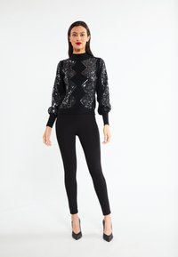 Maglione nero lavorato a maglia con paillettes a motivo diamante, maniche lunghe e silhouette aderente, abbinato a leggings neri e tacchi alti.