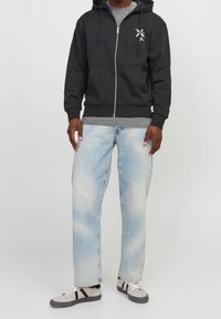Felpa nera con zip e tasche frontali, maglietta grigia sotto, jeans larghi azzurri chiari e sneakers grigie con dettagli neri.
