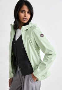 Világos zöld vízálló kabát, kapucnival, amelyen egy barna logós öltözet található az ujjon, fekete mellény és szürke nadrág fölött viselve.