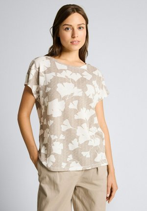 Kobieta ubrana w beżową bluzkę z krótkim rękawem z dużym białym wzorem liści oraz jasnobeżowe spodnie, stojąca z jedną ręką w kieszeni na jednolitym tle.