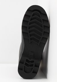 Semelle de botte en caoutchouc noir avec un motif de semelle texturé, présentant de profonds cannelures et une indentation circulaire, le tout sur un fond blanc.