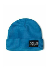 Plava pletena kapa s rebrastom teksturom i preklopljenim rubom. Sadrži crnu oznaku s bijelim natpisom "SURPLUS APPAREL GOODS."
