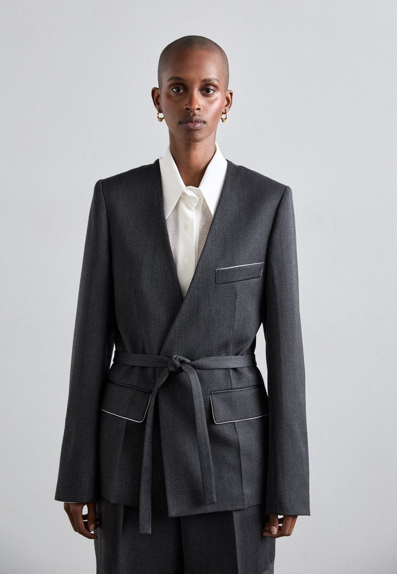 Szürke szabott zakó, melynek stílusa széles, övvel, két elülső zsebbel, fehér díszítéssel, és kivágott gallérral készült, fehér ing felett.