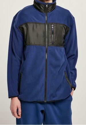 Personne portant une veste en polaire bleue avec une poche poitrine zippée noire et des poches latérales, fermeture éclair frontale complètement fermée.