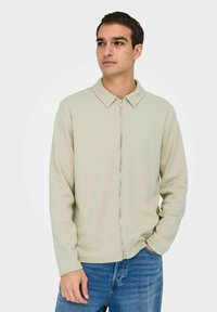 Light beige zip-up jacket with a collar, featuring long sleeves and a smooth texture, paired with blue denim jeans.