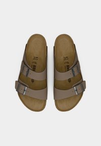 Ζευγάρι καφέ σανδάλια Birkenstock με δύο ρυθμιζόμενες μεταλλικές αγκράφες, πάτο από φελλό και ανοιχτό σχέδιο δαχτύλων πάνω σε λευκό φόντο.