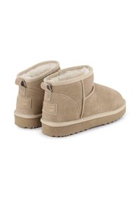 Botas de tobillo de ante beige con un forro suave y esponjoso, diseño minimalista y suela de goma texturizada. Presenta un pequeño lazo en el talón.
