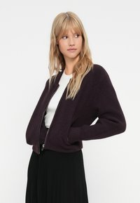 Bjondu sieviete, tērpta tumši violetā zipa džemperī virs balta tops un melnas plisētas svārki, stāv ar rokām kabatās, skatoties pa labi.