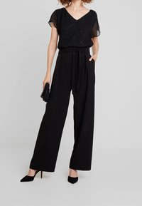 Black sheer blouse with sparkling details, paired with wide-leg black trousers, tied at the waist, and black pointed-toe heels. Accessories included.