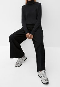 Pull noir à col roulé ; pantalon noir à jambes larges texturé ; chaussures de sport grises et blanches avec semelles noires ; fond minimaliste.