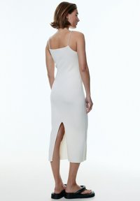 Weißes, geripptes Midi-Kleid mit dünnen Trägern und seitlichem Schlitz, das eine figurbetonte Silhouette bietet. Das Model trägt schwarze Plateau-Latschen.