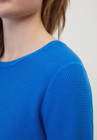 Primo piano di una persona che indossa un maglione di lana blu a maglia, con collo e spalla a vista su uno sfondo neutro.