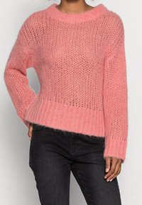 Różowy sweter z dzianiny o luźnym kroju, z szerokim prążkowanym dołem i teksturowanym, otwartym splotem. Posiada długie rękawy i okrągły dekolt.