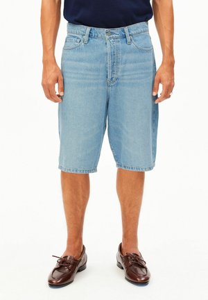 Mann trägt hellblaue Jeansshorts und dunkelbraune Schnürlederschuhe, steht mit locker an den Seiten hängenden Händen.
