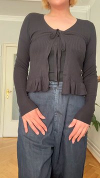 Juodas raštuotas megztinis su gofruota apatine dalimi, su surišamu priekiniu uždarymu, dėvimas ant juodo viršaus ir derinamas su apydančiais tamsiais džinsais.
