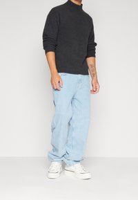 Maglione dolcevita in maglia grigio scuro, vestibilità ampia, abbinato a jeans larghi azzurri chiaro e sneakers bianche con dettagli neri sulle suole.