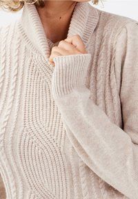 Beiger Strickpullover mit geripptem Kragen und breiten Bündchen. Verziert mit strukturierten Mustern und Zopfmuster-Design. Weicher, gemütlicher Stoff.