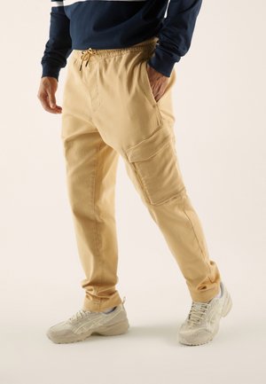 Hombre vistiendo pantalones cargo beige con bolsillos laterales, zapatillas de color beige claro y una camiseta de manga larga azul marino con rayas blancas.