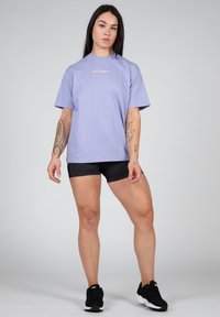 Light purple cotton t-shirt with short sleeves and a round neckline; features small orange logo on the chest; paired with black athletic shorts.