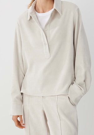 Frau trägt cremfarbenes Cord-Hemd mit langen Ärmeln und Kragen sowie passende hoch taillierte Hose, Hand lässig in der Tasche, vor einfachem Hintergrund.