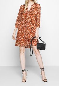Femme portant une robe orange à motifs longueur genou, des talons noirs à brides, et tenant un petit sac à main noir avec une longue bandoulière.