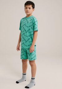 Turkizni pižamo set s črnimi napisnimi vzorci, kratkimi rokavi in kratkimi hlačami, v kombinaciji s sivo obutvijo, ki ima črne črte na vrhu.