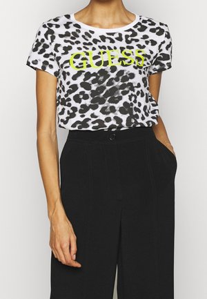 Vrouw draagt een wit en zwart luipaardprint t-shirt met gele "GUESS" tekst en zwarte high-waisted broek, één hand in de zak.