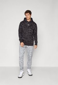 Férfi fekete mintás kapucnis felsőben és szürke mintás melegítőnadrágban, fehér tornacipővel, egyszínű világosszürke háttér előtt állva.