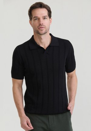 Rövid barna hajú férfi, fekete, rövid ujjú, texturált galléros pólót és sötétzöld nadrágot visel, világos háttér előtt áll.