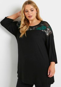 Schwarzes Tunika-Oberteil mit Dreiviertelärmeln, versehen mit transparenten Schulterpaneelen und kunstvollen floralen Stickereien in Grün und Silber. Glattes Material.