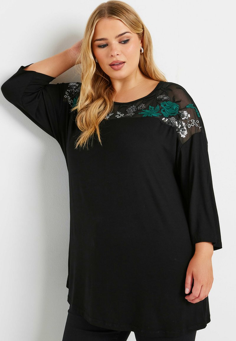Schwarzes Tunika-Oberteil mit Dreiviertelärmeln, versehen mit transparenten Schulterpaneelen und kunstvollen floralen Stickereien in Grün und Silber. Glattes Material.