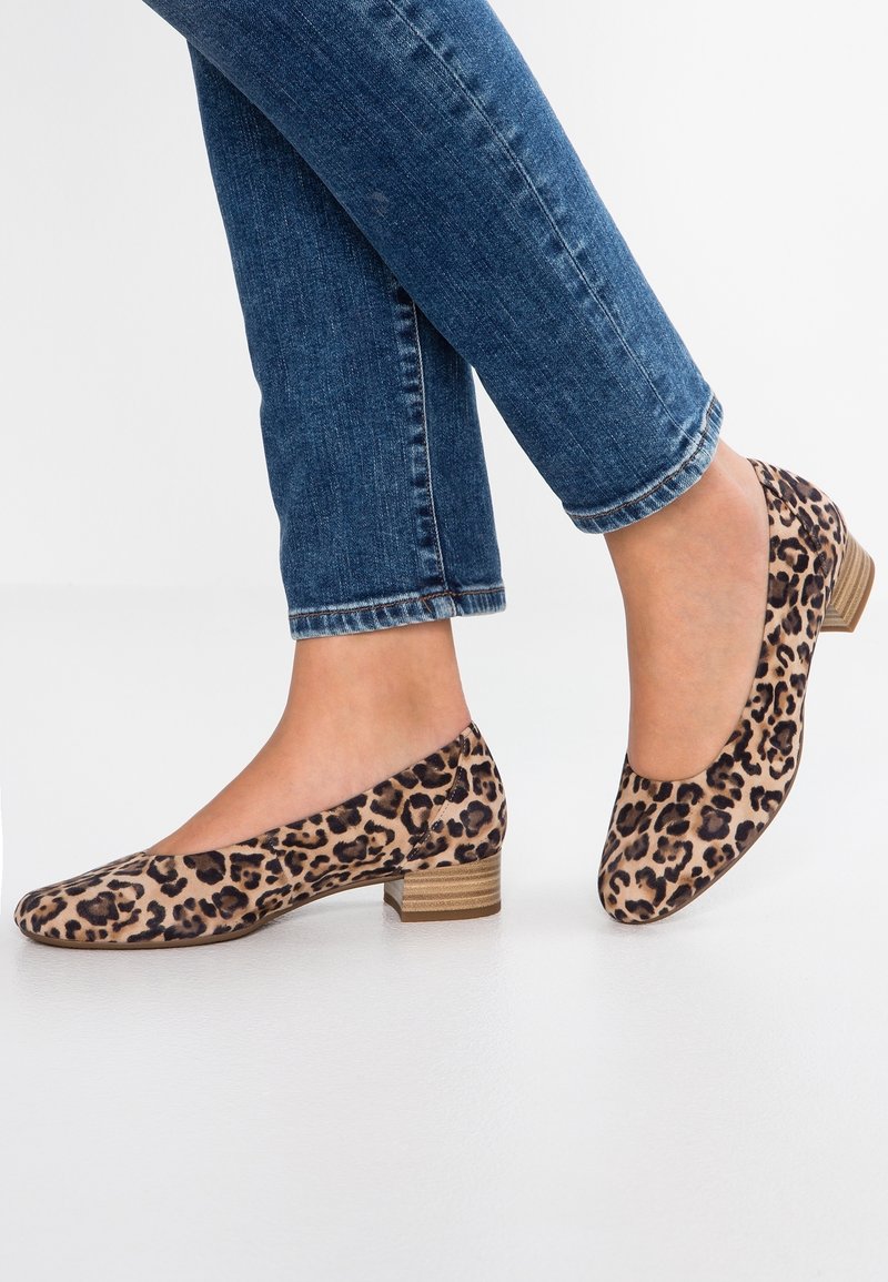 Personne portant un jean bleu et des chaussures à talons bas imprimé léopard, debout avec un pied légèrement levé sur un sol blanc.