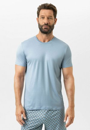 Homme barbu portant un t-shirt bleu clair à manches courtes et un short bleu à motifs, debout devant un fond blanc uni.