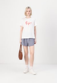 Vit T-shirt med en färgglad virveldesign, blå och vita randiga shorts, vita sneakers och en brun axelväska.
