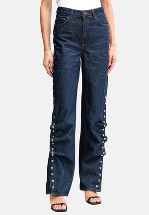 Vrouw draagt donkerblauwe spijkerbroek met hoge taille en zilveren drukkers langs de buitennaad van de pijpen, gecombineerd met zwarte open teen sandalen.